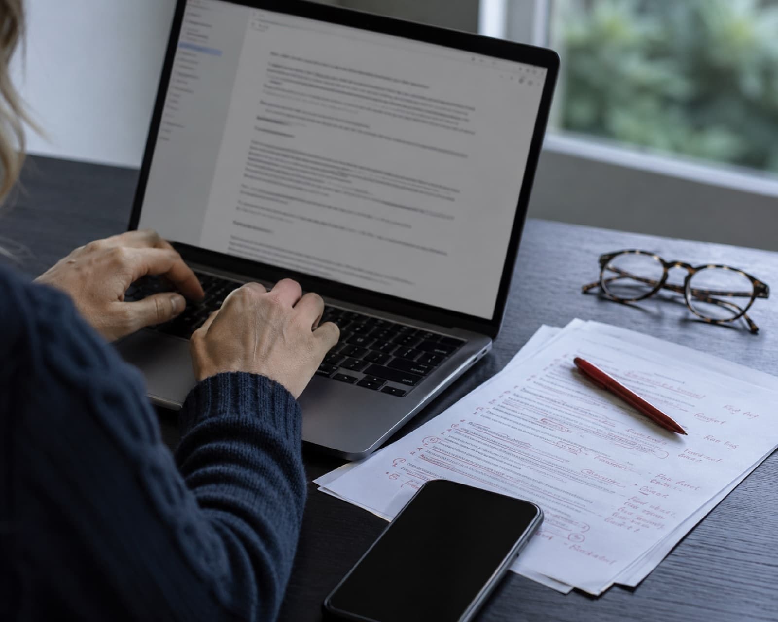 A writer editing a fundraising email on a laptop beside a printed draft marked up in red pen.